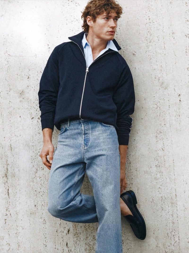 Valentin Humbroich wears a dark zip-up knit jacket over a pale blue collared shirt with mid-wash jeans and dark leather loafers against a concrete wall
