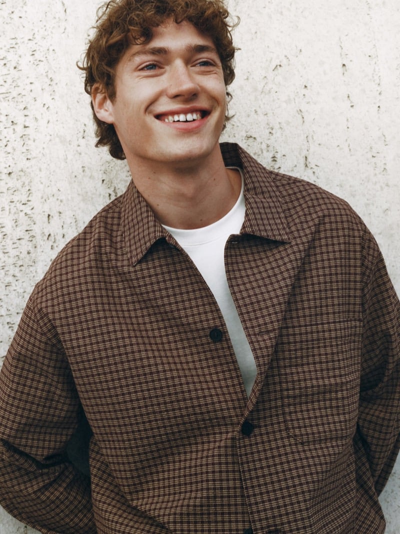 Smiling portrait of Valentin Humbroich in a brown micro-check overshirt layered over a plain tee against a pale concrete wall
