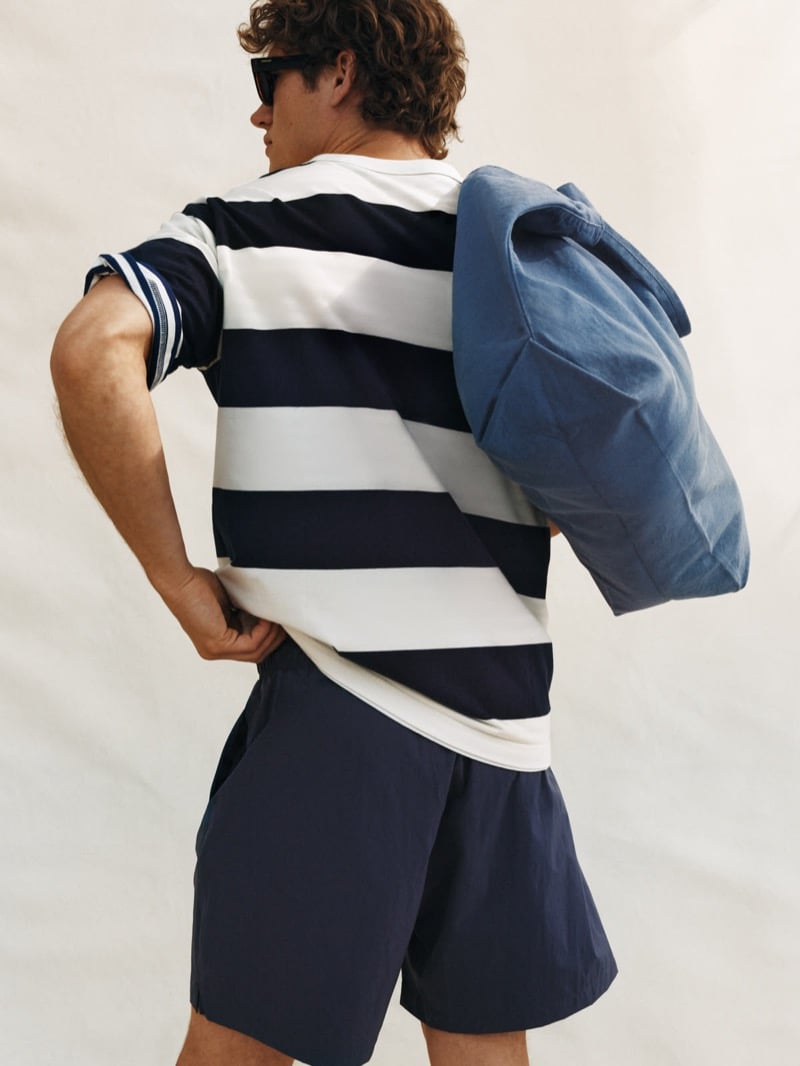Valentin Humbroich seen from behind in dark sunglasses, wearing a bold rugby-striped short-sleeve tee with dark drawstring shorts and a washed blue canvas tote slung over one shoulder