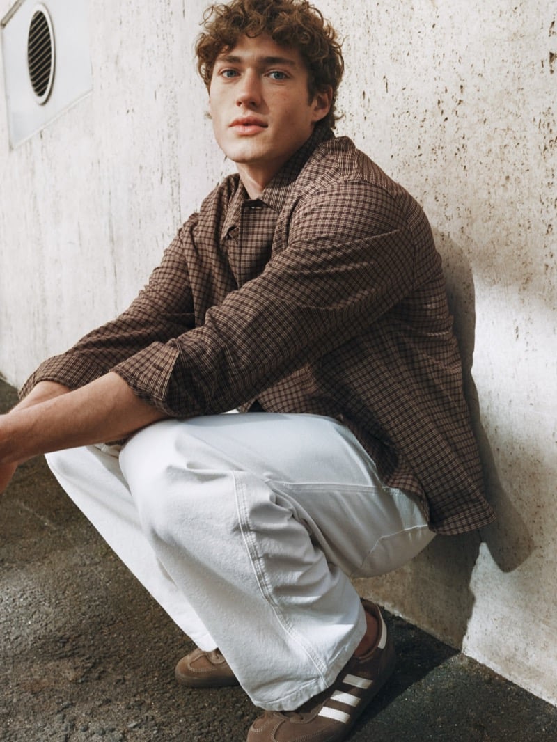 Valentin Humbroich crouches against a concrete wall in a brown micro-check overshirt with pale wide-leg jeans and brown three-stripe suede sneakers