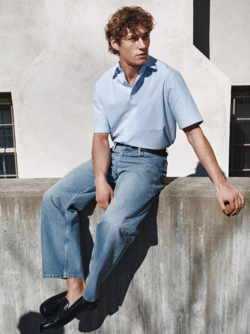 Valentin Humbroich sits on a concrete wall in a pale blue short-sleeve shirt, mid-wash jeans, a braided belt, and dark leather loafers