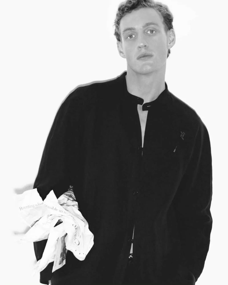 Black and white portrait of Aubrey O'Mahony in a black mandarin-collar button-up shirt by Giorgio Armani, holding a crumpled sheet of newsprint at his hip