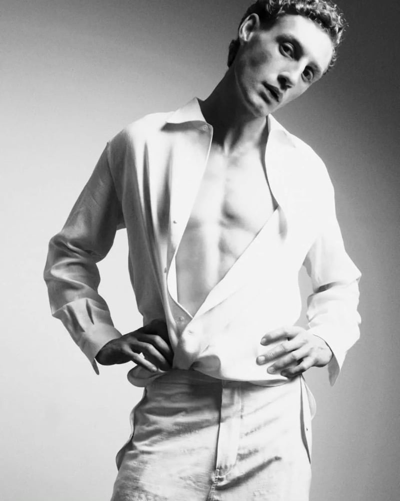 Black and white portrait of Aubrey O'Mahony tugging at the waist of a white linen shirt left unbuttoned over a bare chest and pale white trousers