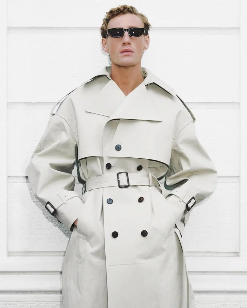 Aubrey O'Mahony stands against a paneled white wall in a Givenchy double-breasted belted trench coat with hands in the pockets and small black rectangular sunglasses