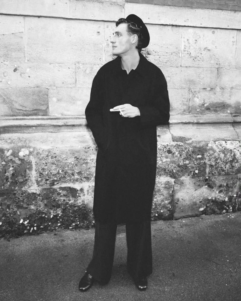 Black and white image of Aubrey O'Mahony leaning against a Parisian stone wall in a long black overcoat with wide-leg black trousers, leather loafers, and a black beret, a lit cigarette in one hand