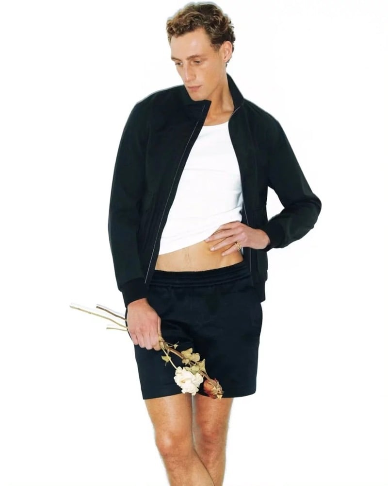 Aubrey O'Mahony wears a black bomber jacket over a white ribbed tank with matching black drawstring shorts, holding a bundle of dried white and pink roses against a white backdrop