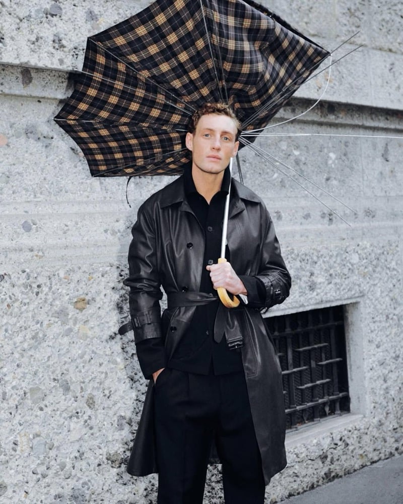 Aubrey O'Mahony leans against a stone wall in a black leather belted trench by Brioni over a black button-up and trousers, lifting a brown and black plaid umbrella that has blown inside out above his head