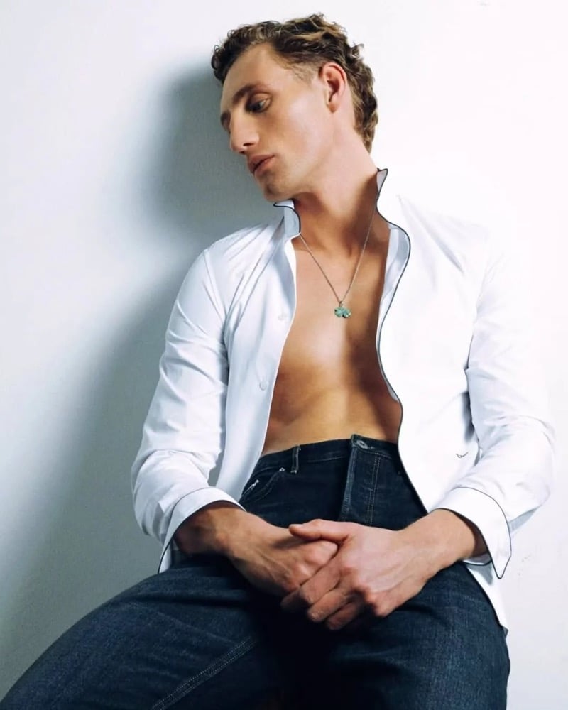 Aubrey O'Mahony leans against a white wall in an open white shirt with navy piping and dark blue jeans, a turquoise gemstone pendant resting on his bare chest