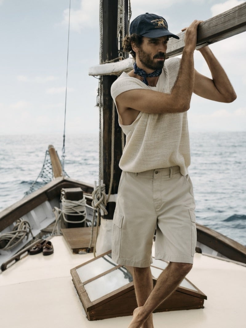 Nuno Marques shoulders the boom of a sailboat at sea, wearing a ribbed knit vest, archive-reissue cargo shorts, a blue bandana, and a Banana Republic logo cap