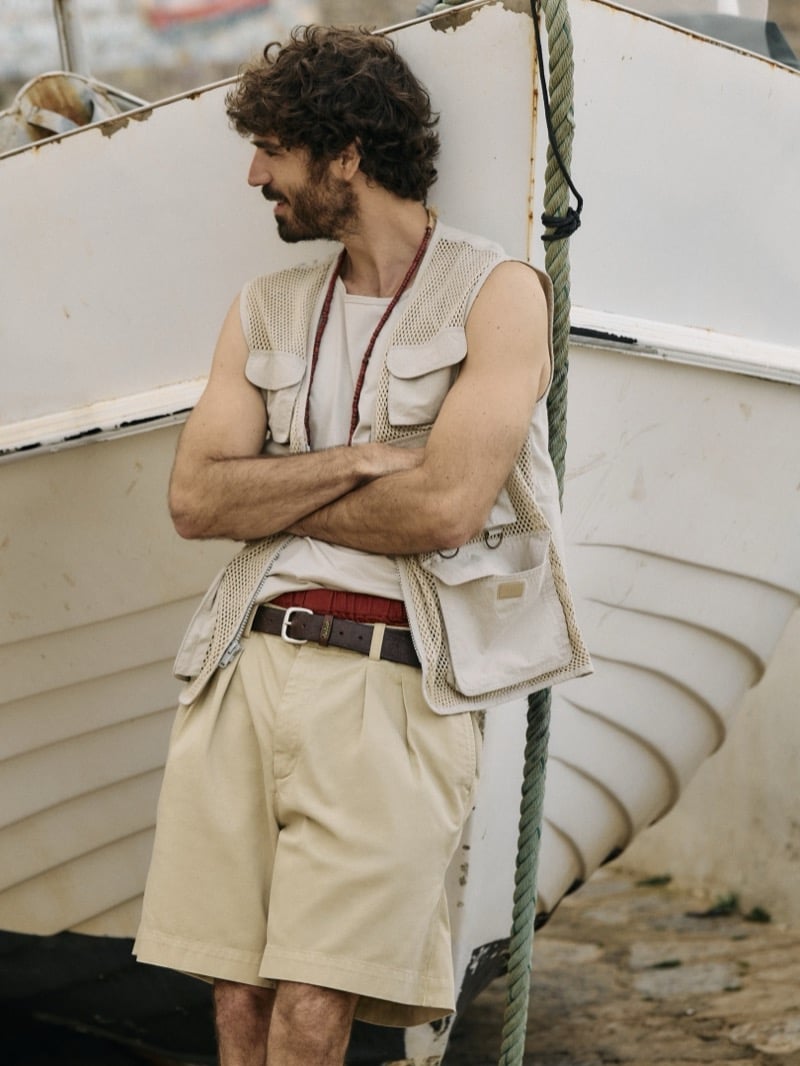 Nuno Marques leans in profile against a beached fishing boat in a mesh photojournalist utility vest layered over a ribbed tank, a leather belt, and pleated shorts