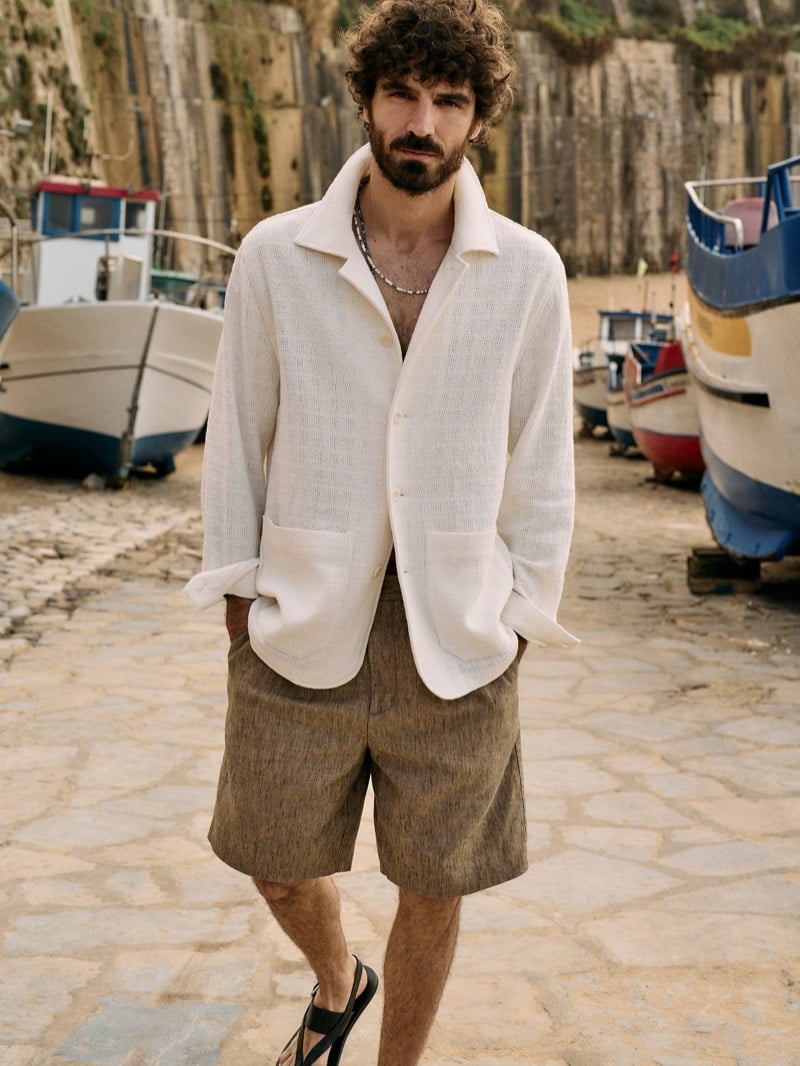 Nuno Marques stands on a cobblestone harbor walk in an open basketweave linen shirt jacket, earth-toned linen Bermuda shorts, a beaded necklace, and leather sandals with fishing boats behind him