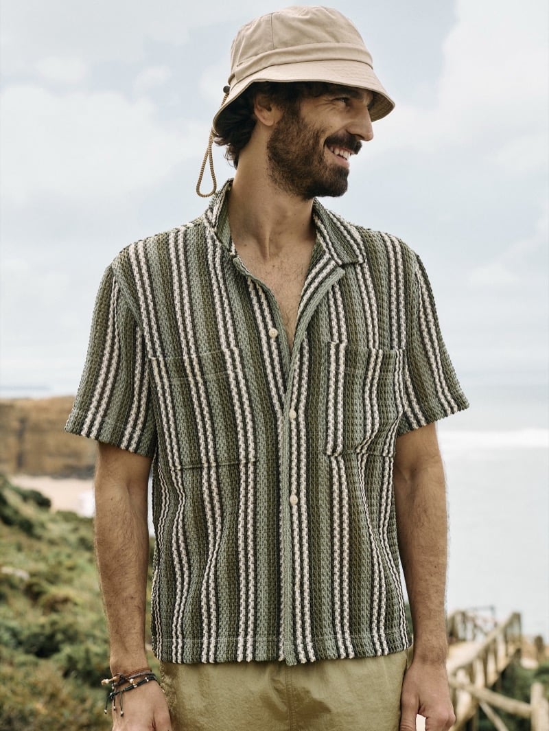 Nuno Marques smiles in a green crochet-stripe camp collar resort shirt and a drawstring bucket hat against the Atlantic coastline
