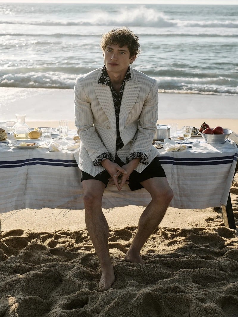 Valentin Humbroich seated on the edge of a long beachside table set for lunch in a cream Italian cotton-linen seersucker double-breasted suit jacket layered over a black and cream geometric-print shirt with black shorts, barefoot on the sand as Atlantic waves break behind him