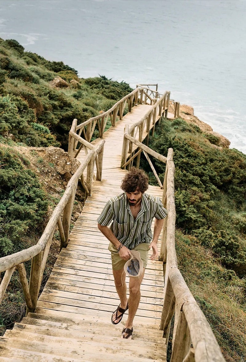 Nuno Marques climbs a wooden cliff-side boardwalk in a green crochet-stripe camp shirt, olive shorts, and leather fisherman sandals while holding a bucket hat