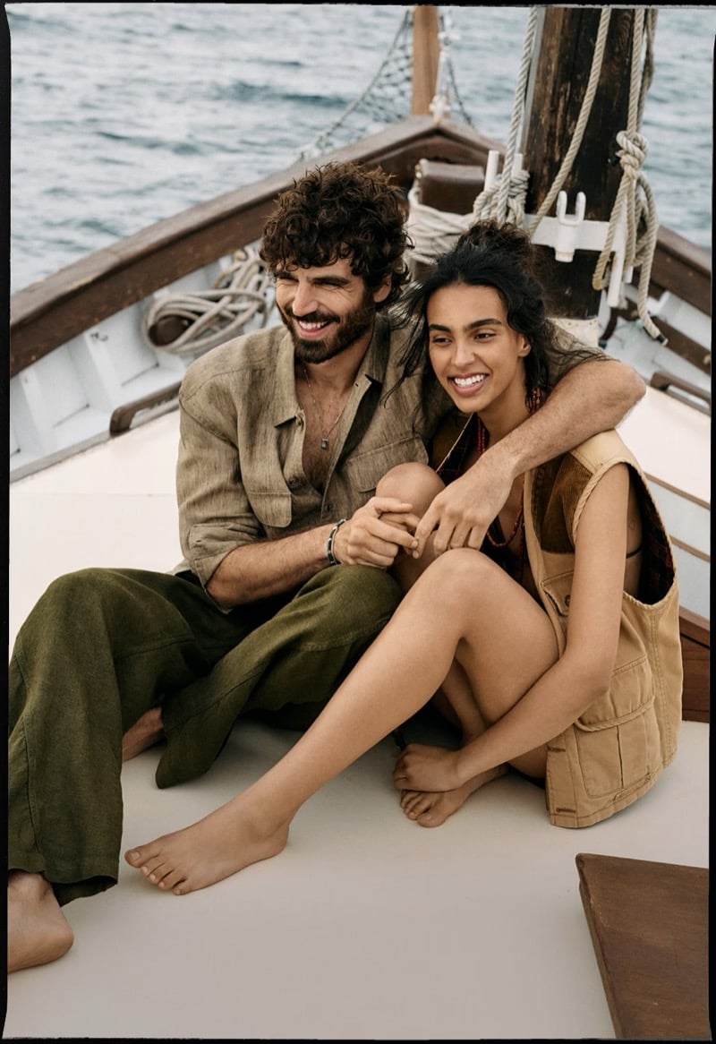 Nuno Marques and a female model laugh seated on a wooden boat deck, Nuno in an open linen camp shirt and olive linen beach pants and his companion in a Banana Republic cargo utility vest
