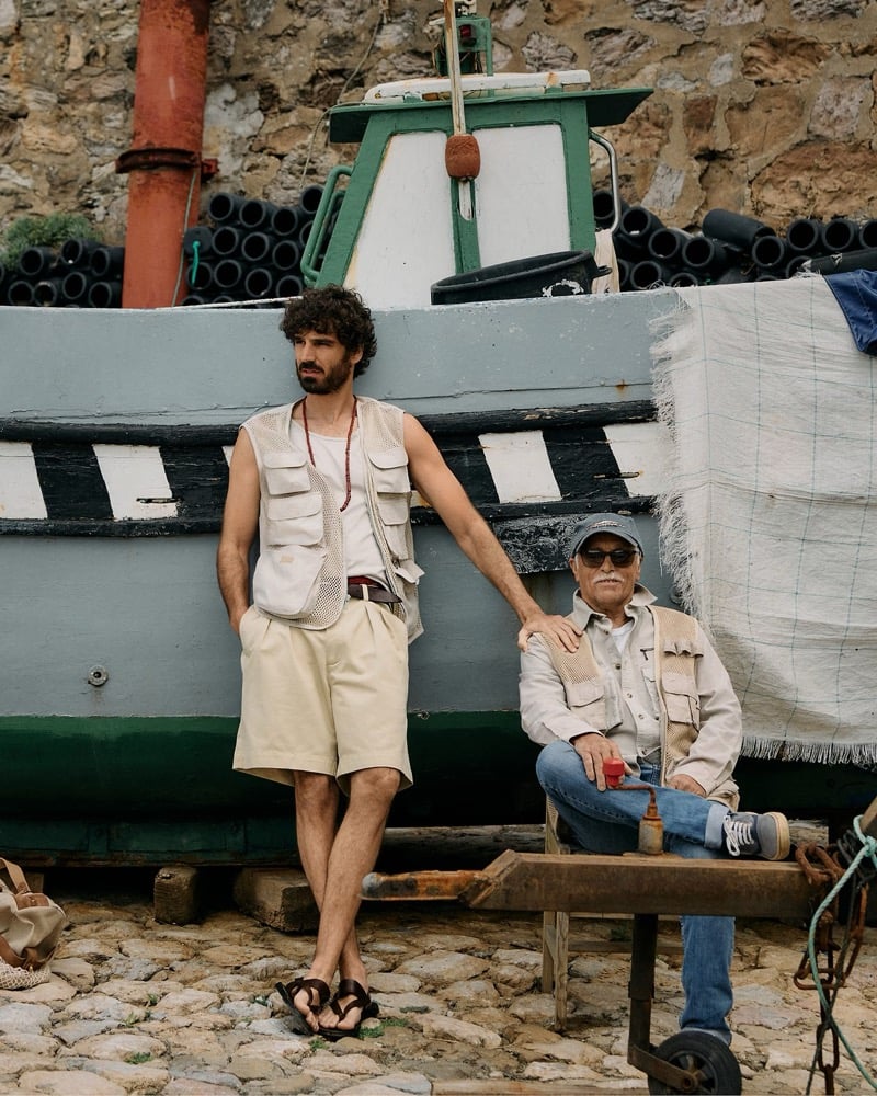 Nuno Marques stands alongside a seated older Ericeira fisherman against a fishing boat hull, Nuno wearing the mesh photojournalist vest, ribbed tank, pleated shorts, and leather sandals