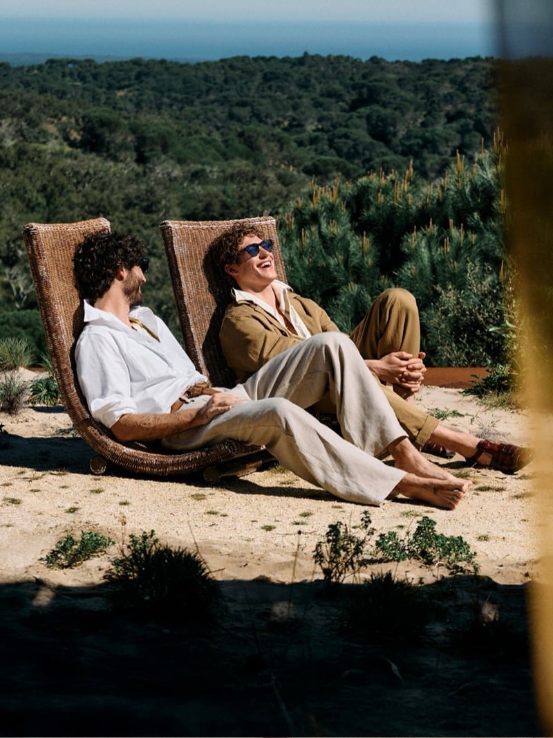 Nuno Marques and Valentin Humbroich reclining in twin woven wicker lounge chairs on a sandy clifftop above pine-covered Alentejo hills, the Atlantic visible in the distance, Nuno in a white shirt and beige linen trousers, Valentin in a tan overshirt over a cream tee with cream linen trousers