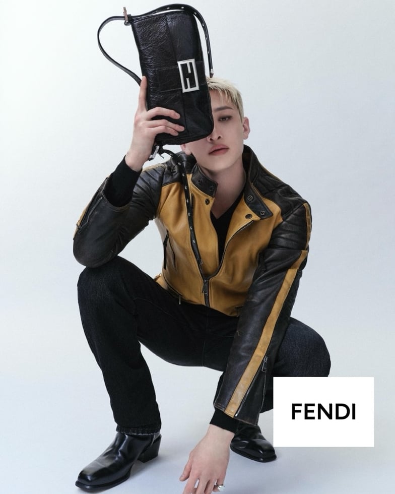 Bang Chan crouches in a yellow and black colorblocked leather biker jacket, dark denim, and square-toe leather boots, lifting the elongated Fendi Baguette 26424 in crinkled black leather across his face
