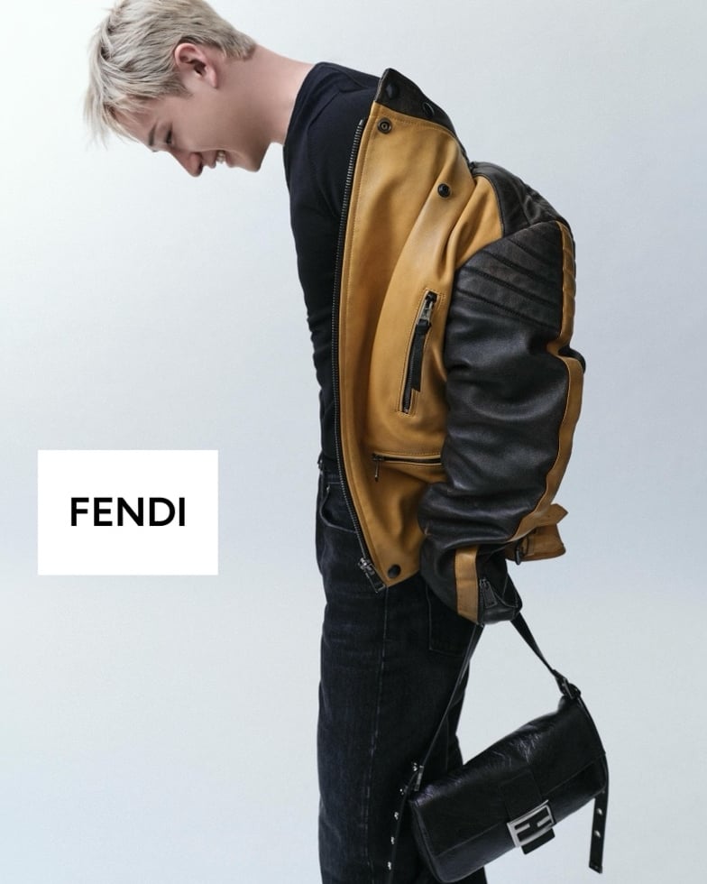 Bang Chan turns in profile with head bowed, the yellow and black leather biker jacket worn over a fitted black top, the elongated Fendi Baguette 26424 in crinkled black leather hanging from a slim shoulder strap at his hip