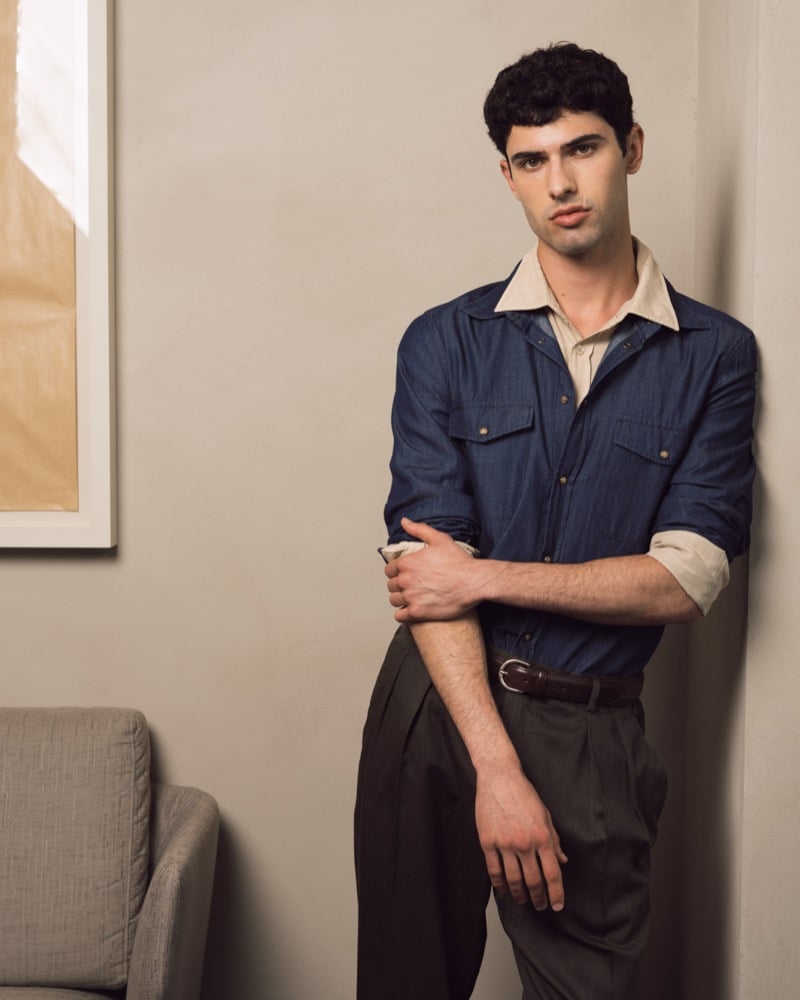 Model Alessandro Rengo leans against a beige wall in a navy denim western shirt layered over a cream collared shirt, paired with pleated dark brown trousers and a brown leather belt