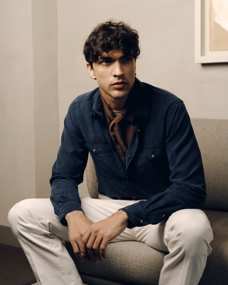 Model Yiorgos Paraskeva sits on a beige sofa in a navy linen utility shirt with chest pockets, a brown neckerchief over a cream T-shirt, and cream cotton trousers