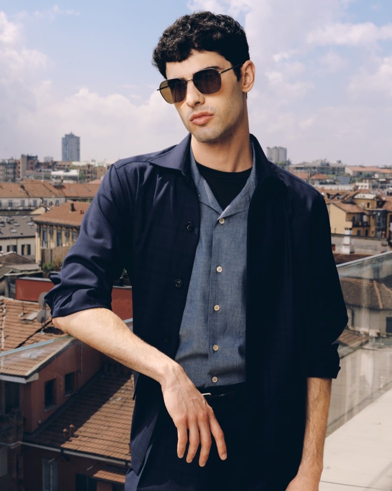 Model Alessandro Rengo wears aviator sunglasses, a navy overshirt over a chambray shirt and black T-shirt, on a Milan rooftop with the city skyline behind him