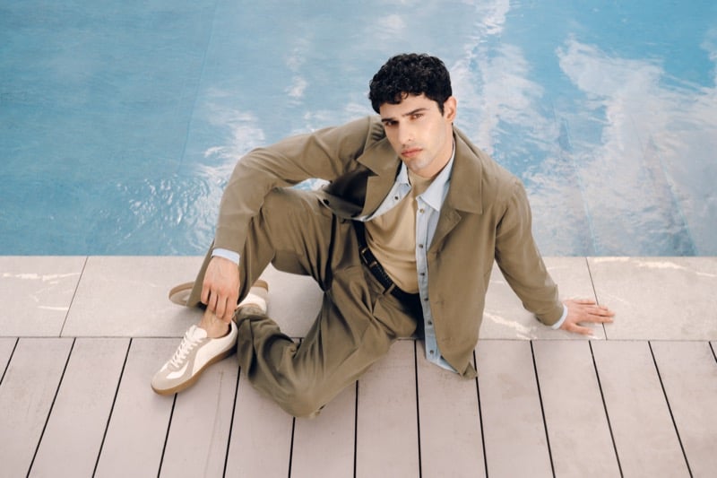 Model Alessandro Rengo sits poolside in a tobacco jacket and matching trousers over a pale blue shirt, paired with cream low-top sneakers