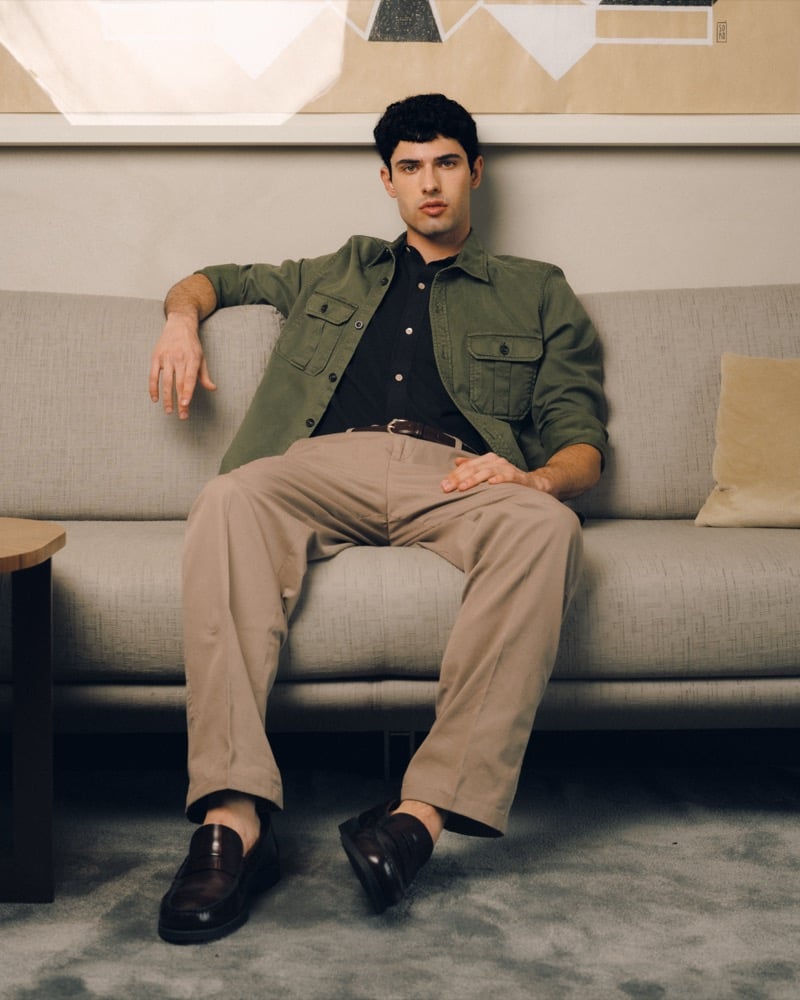 Model Alessandro Rengo on a gray sofa in an olive cotton overshirt with chest pockets over a navy shirt, paired with stone cotton trousers, a brown leather belt, and dark brown penny loafers