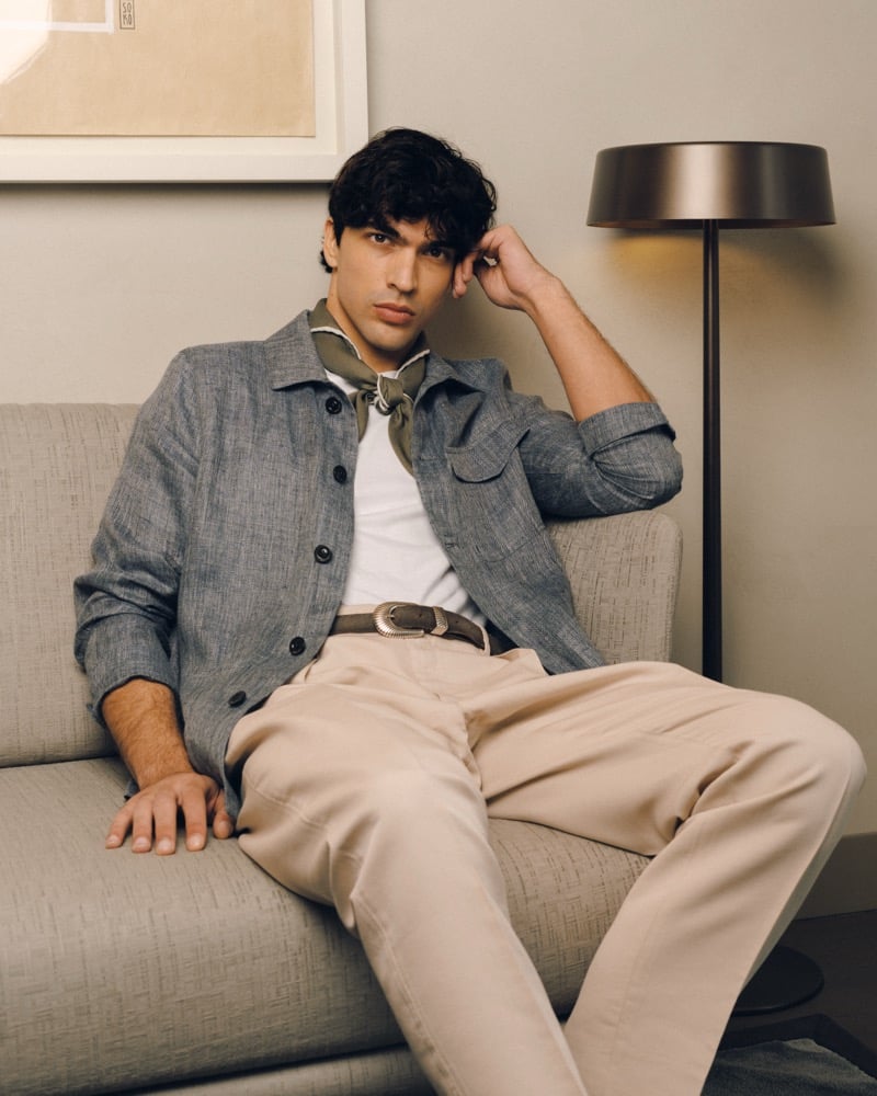 Model Yiorgos Paraskeva seated on a sofa in a chambray cotton overshirt with chest pocket over a white T-shirt, an olive neckerchief, and cream high-waisted trousers with a sculpted silver belt buckle