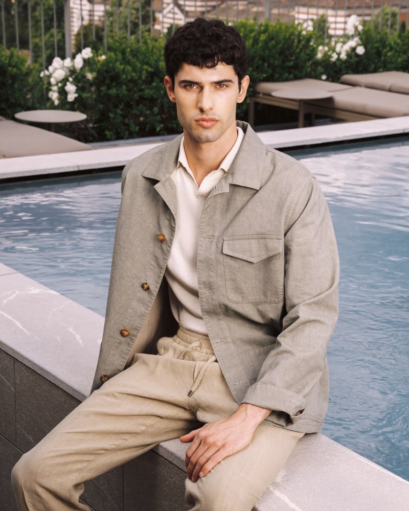 Model Alessandro Rengo sits poolside in a stone linen utility jacket open over a cream knit polo, paired with sand drawstring trousers
