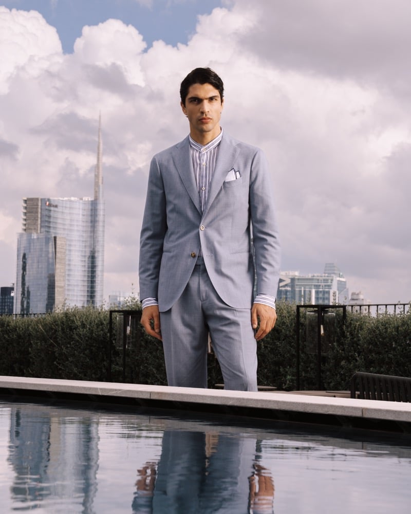 Model Yiorgos Paraskeva wears a dusty blue micro-check suit with a soft single-breasted jacket and pleated trousers over a navy and white striped shirt with a white pocket square, photographed by a rooftop pool with the Milan skyline behind him