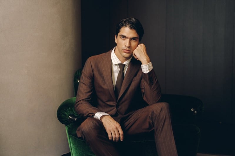 Model Yiorgos Paraskeva sits on a green velvet chair in a tobacco pinstripe double-breasted suit with peak lapels, paired with a cream shirt and brown silk tie