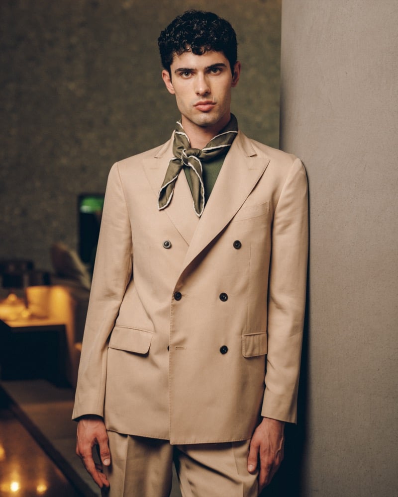 Model Alessandro Rengo in a stone soft double-breasted suit with peak lapels, layered over an olive knit polo and an olive printed neckerchief