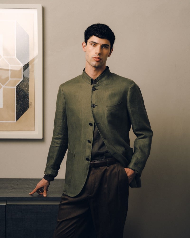 Model Alessandro Rengo in an olive linen collarless jacket with patch pockets, layered over a darker olive shirt and worn with brown pleated trousers
