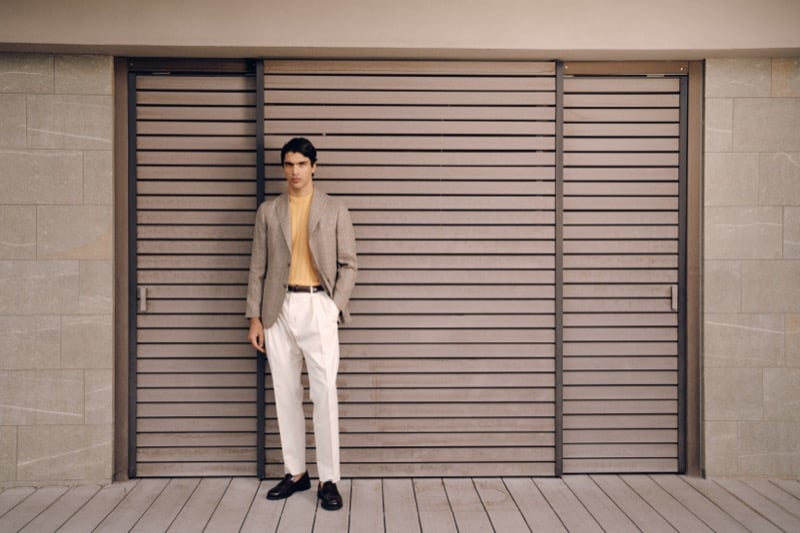 Model Yiorgos Paraskeva in a tan checked linen blazer over a yellow knit polo, paired with cream pleated trousers, a brown belt, and dark loafers