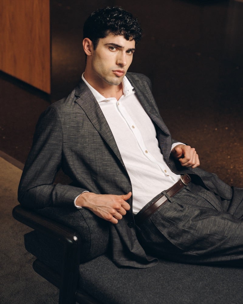 Model Alessandro Rengo reclines in a charcoal linen soft suit with a single-breasted notch lapel jacket and wide pleated trousers, paired with a white shirt and brown leather belt