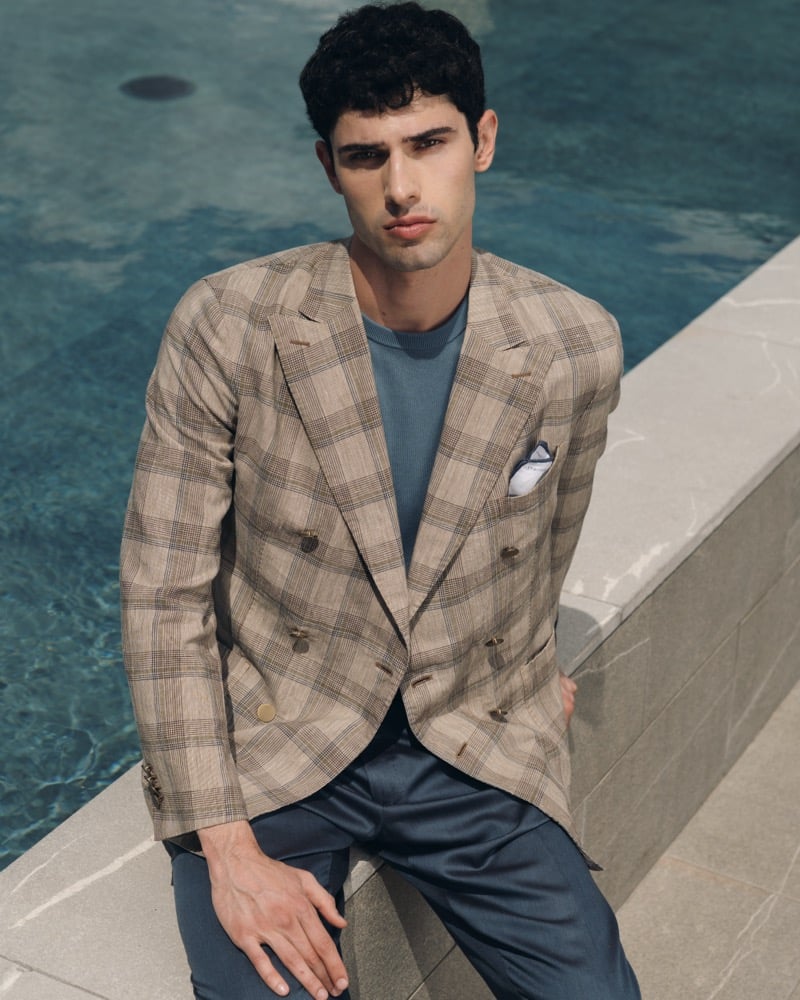 Model Alessandro Rengo poolside in a tan Prince of Wales check double-breasted blazer with peak lapels over a dusty blue crewneck and matching blue trousers, finished with a white pocket square