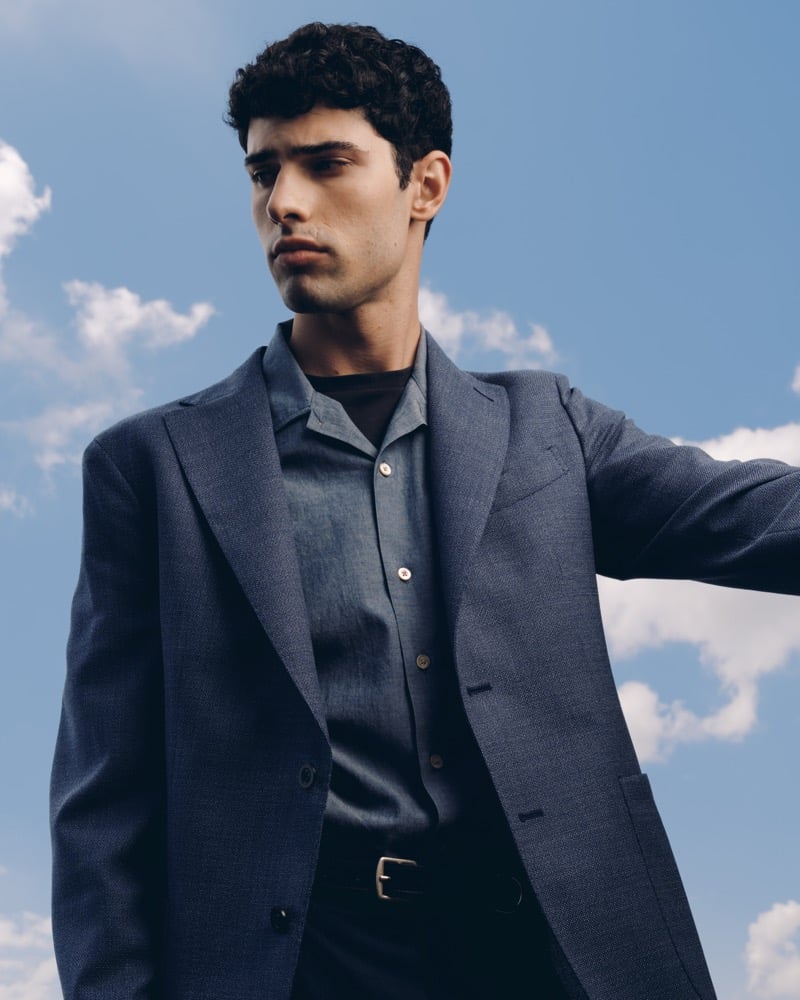 Model Alessandro Rengo photographed against a blue sky in a dusty blue soft single-breasted blazer over a chambray shirt and black T-shirt with black trousers
