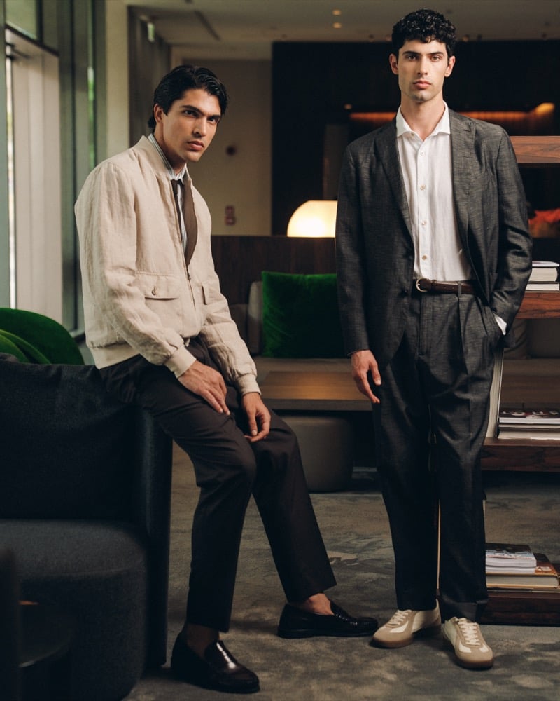 Models Yiorgos Paraskeva and Alessandro Rengo — Yiorgos in a stone linen bomber jacket with chest pockets, a brown striped tie, and dark brown trousers with loafers, Alessandro in a charcoal linen suit with a white shirt, brown belt, and stone low-top sneakers