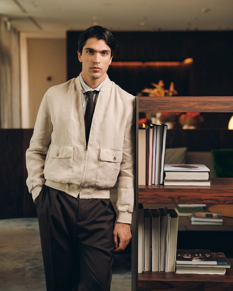 Model Yiorgos Paraskeva in a stone linen bomber jacket with chest pockets over a striped shirt and brown knit tie, paired with dark brown pleated trousers