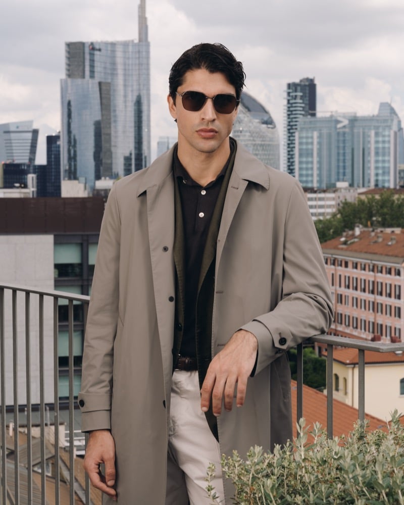 Model Alessandro Rengo on a Milan rooftop wearing aviator sunglasses, a stone lightweight trench coat layered over an olive blazer, a black polo shirt, and cream trousers