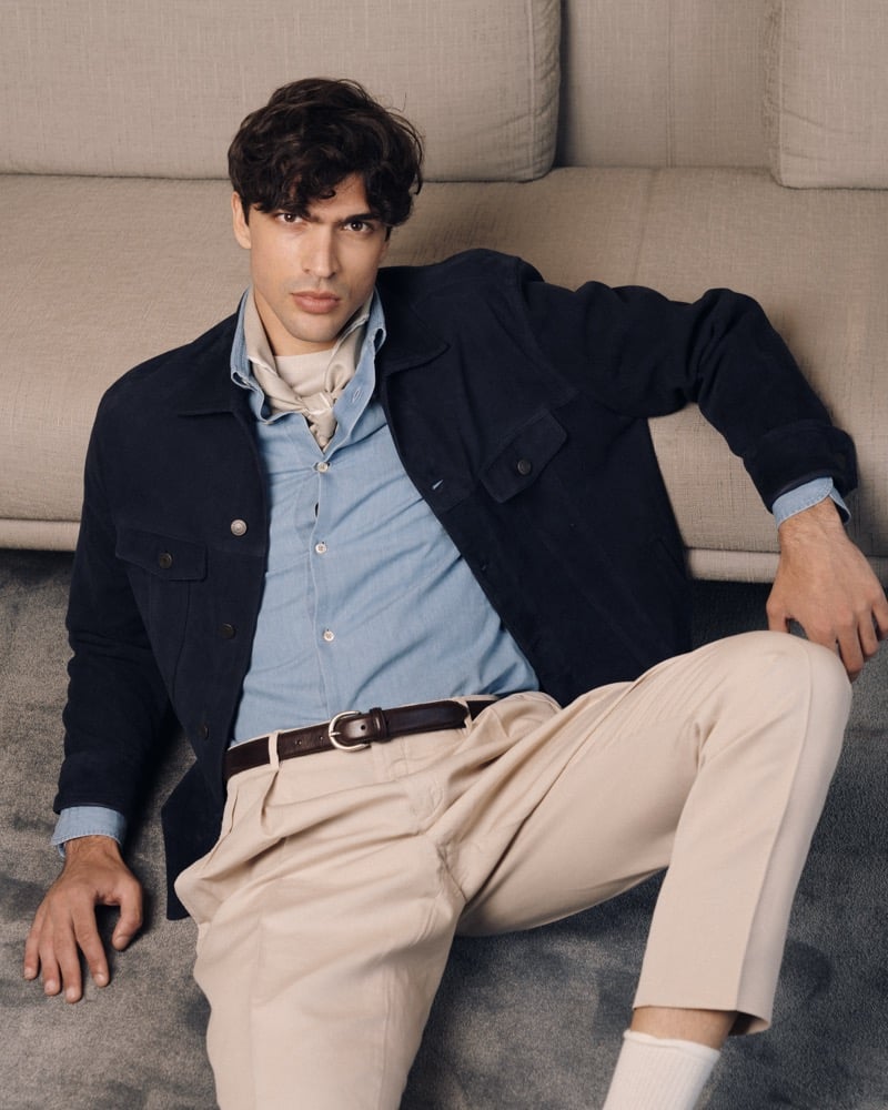 Model Yiorgos Paraskeva reclines on a beige rug against a sofa in a navy suede trucker jacket over a chambray shirt and cream neckerchief, paired with high-waisted stone pleated trousers and a brown belt