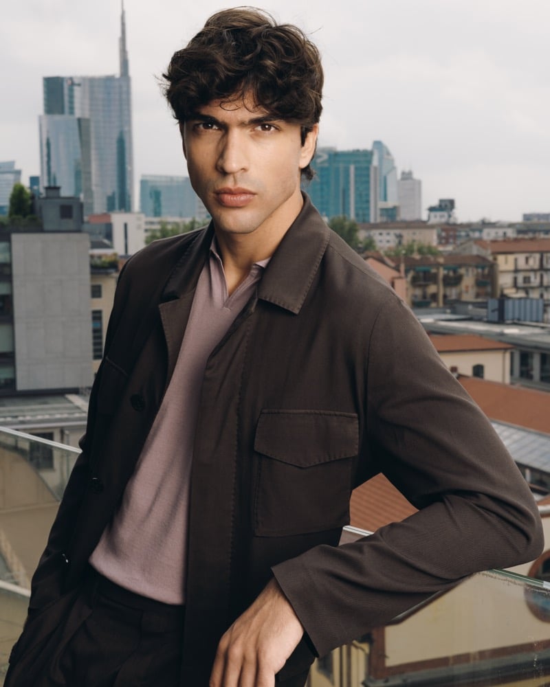 Model Yiorgos Paraskeva on a Milan rooftop in a dark brown cotton overshirt with chest pocket layered over a faded lavender knit polo, with the city skyline behind him