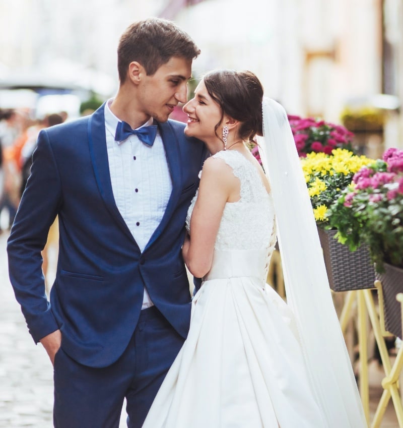 Groom in a blue shawl-lapel tuxedo and matching bow tie standing forehead to forehead with his bride in a white lace ball gown and a long veil, on a European street lined with flower boxes