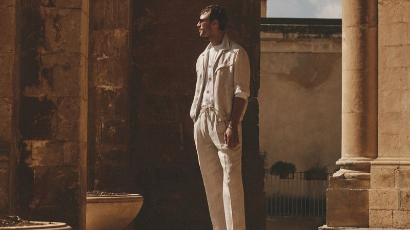 Clément Chabernaud stands between Sicilian stone columns in a stone linen utility shirt-jacket and drawstring trousers, looking out toward the courtyard