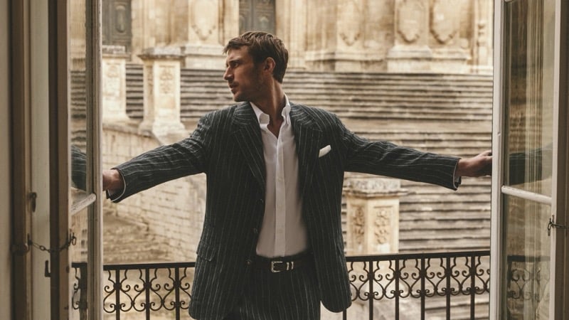 Clément Chabernaud stretches his arms across the open French doors of a Modica balcony in a charcoal pinstripe linen suit over a white shirt