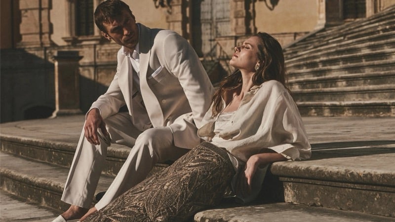 Clément Chabernaud in a stone double-breasted suit sits on baroque stone steps beside a female model in a sheer ivory blouse and metallic embellished skirt