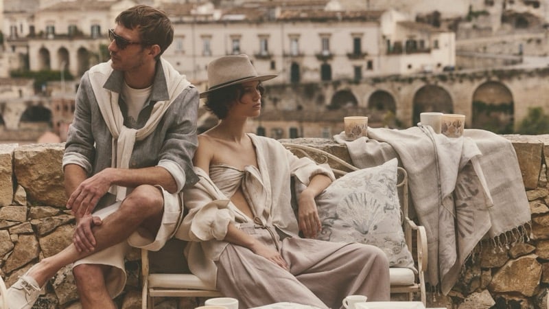 Clément Chabernaud in linen shorts and a tossed-on shirt sits on a terrace overlooking Modica, beside a female model in stone separates and a tan brimmed hat