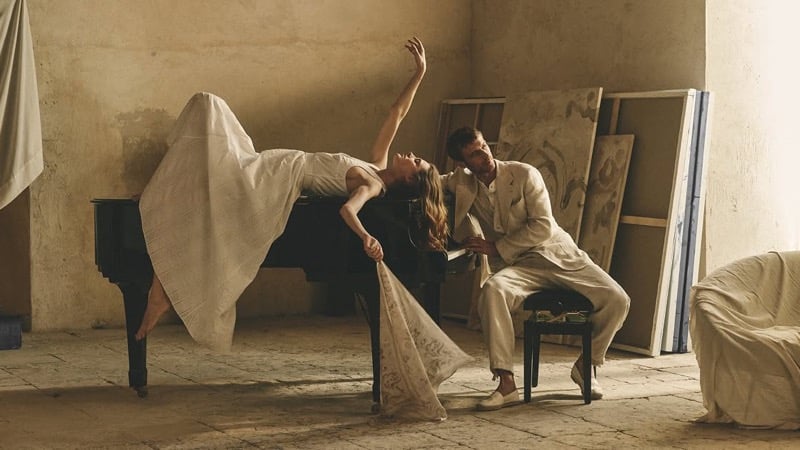 Clément Chabernaud at the piano in a stone linen suit while a female model in a long ivory gown stretches across the top of the instrument inside a Sicilian artist studio