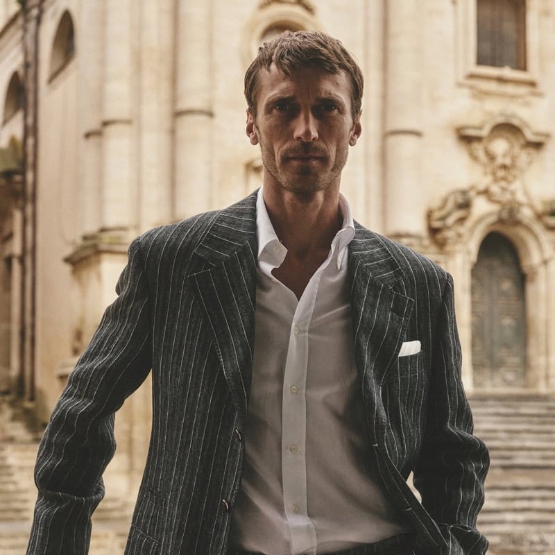 Clément Chabernaud walks through a Modica piazza in a charcoal pinstripe linen blazer worn open over a white poplin shirt with a folded pocket square, baroque limestone facade behind him